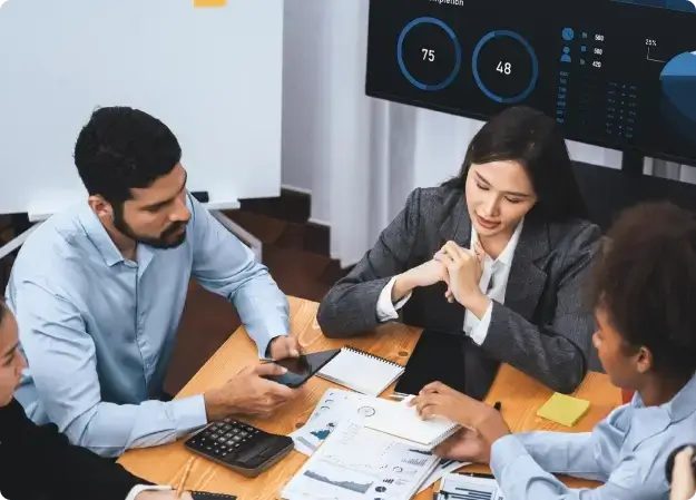 Four colleagues around a table reviewing charts and notes, a dashboard screen behind them